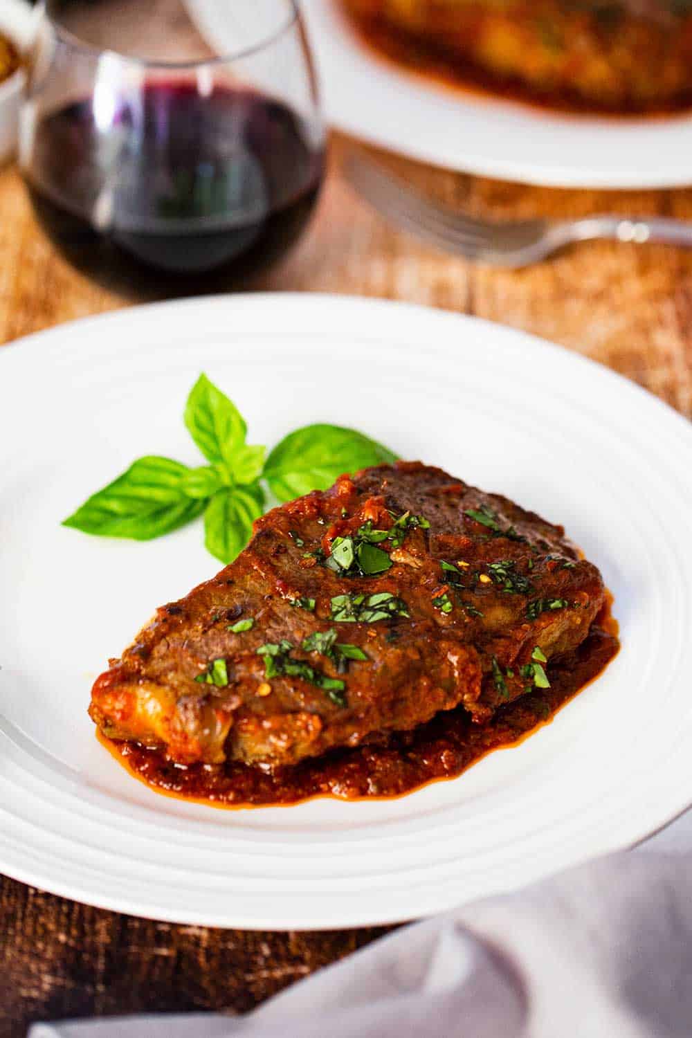 A single serving of steak pizzaiola on a white dinner plate sitting next to a glass of red wine.