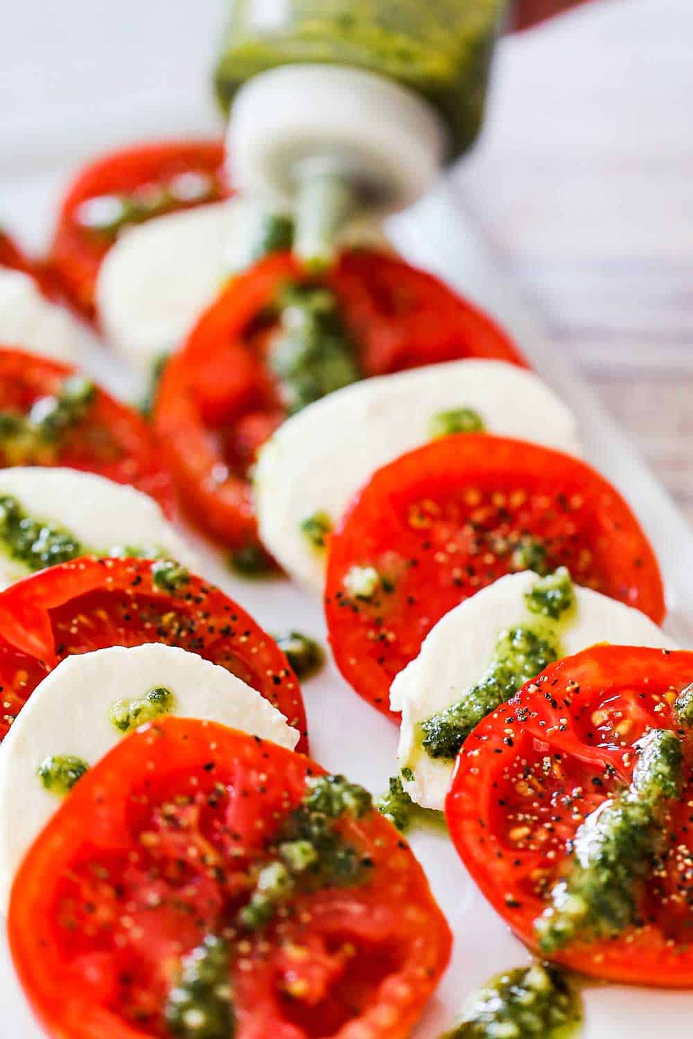 A white rectangular platter holding layers of slice tomatoes and mozzarella to form a caprese salad being topped with squirts of pesto sauce from a squeeze bottle.