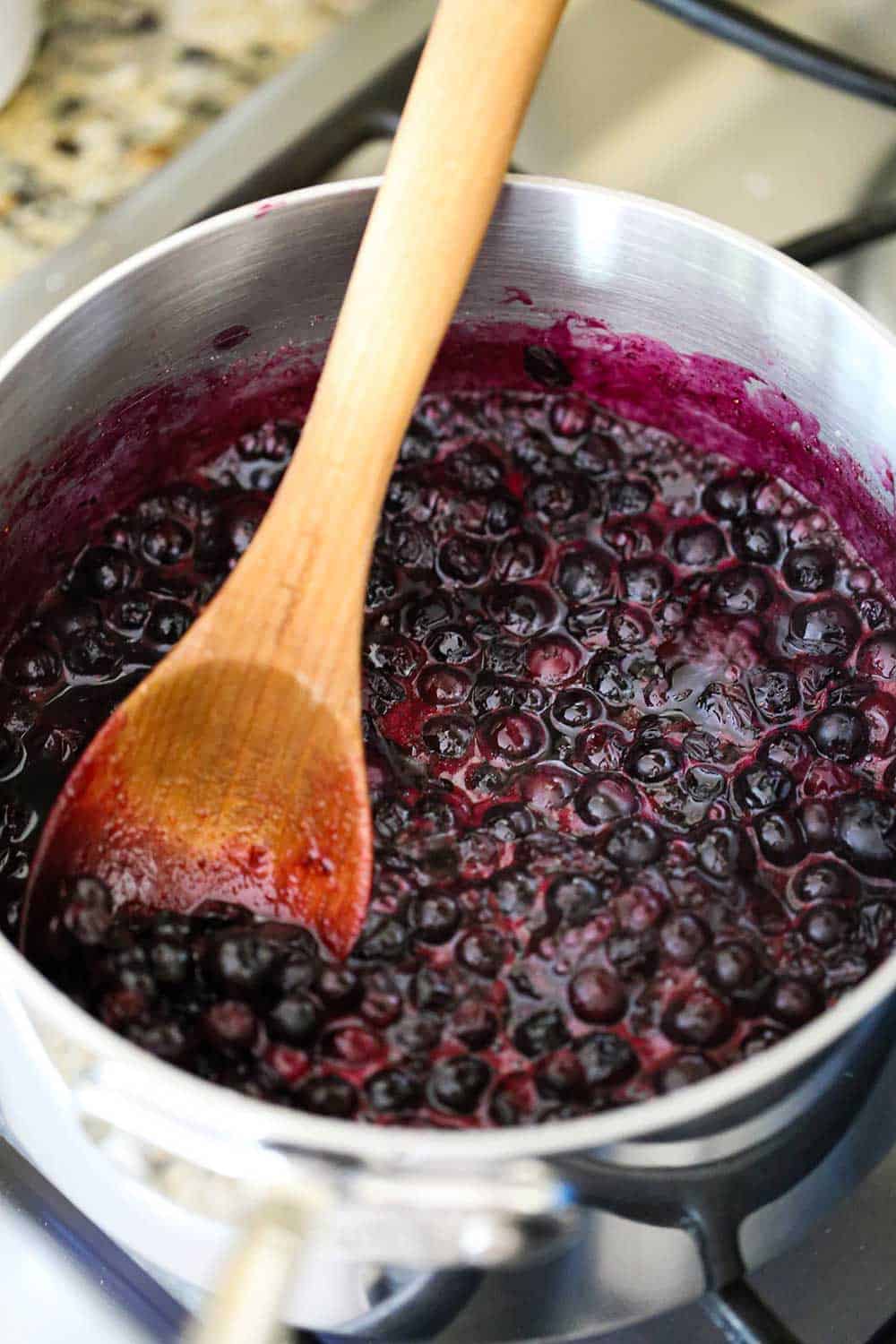 A silver saucepan filled with simmering blueberries with a wooden spoon stirring them all.
