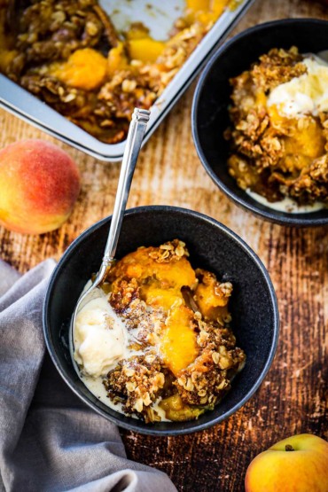 Two blue dessert bowls filled with peach crisp with a dollop of melting vanilla ice cream off to the side all next to a couple of peaches and a pan of the crisp.