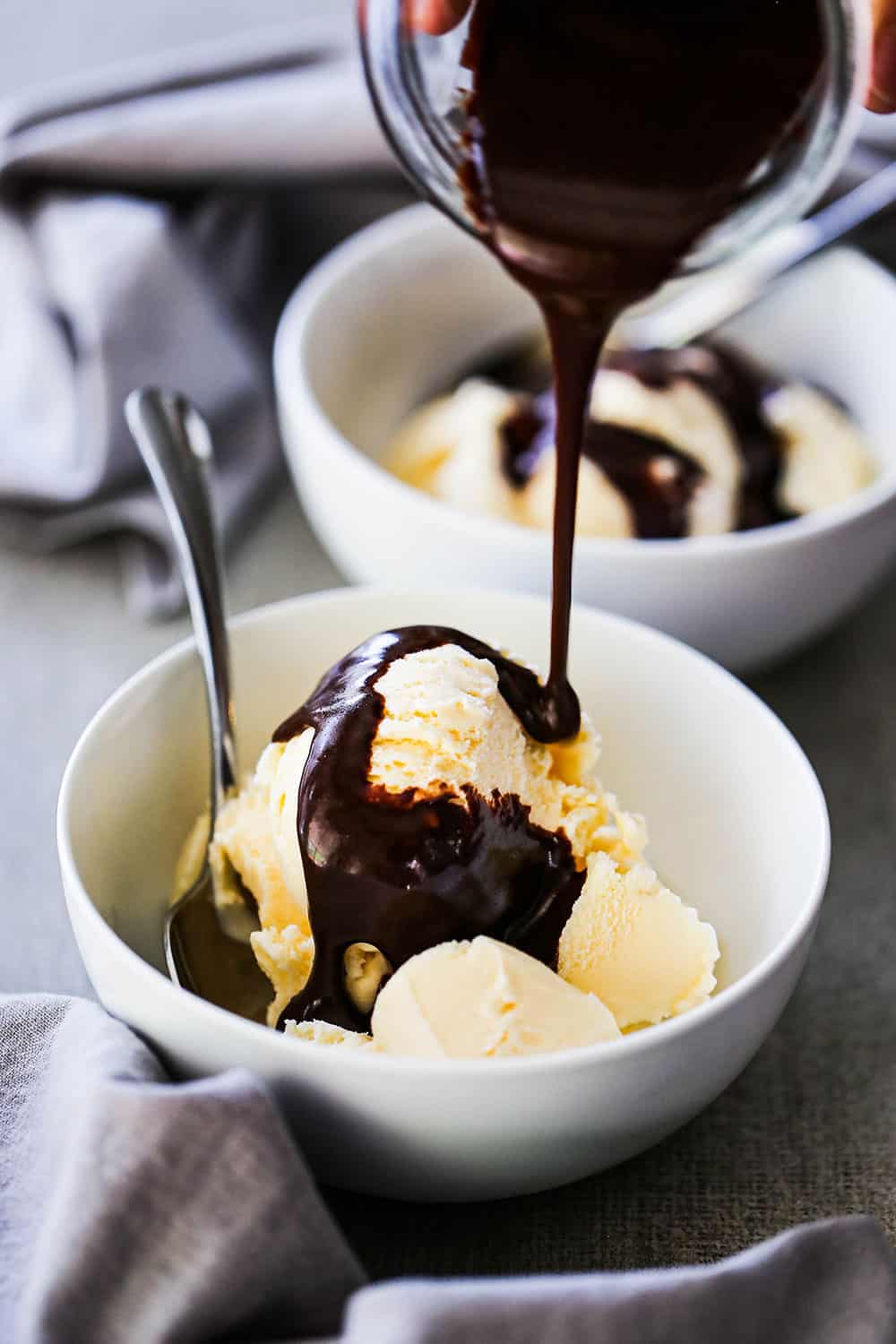 A small jar of homemade chocolate sauce being poured over a bowl filled with several scoops of vanilla ice cream.