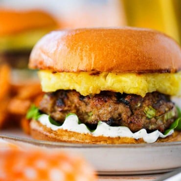 A curry turkey burger sitting on green leaf lettuce and dill yogurt sauce topped with a grilled pineapple slice in a brioche bun.