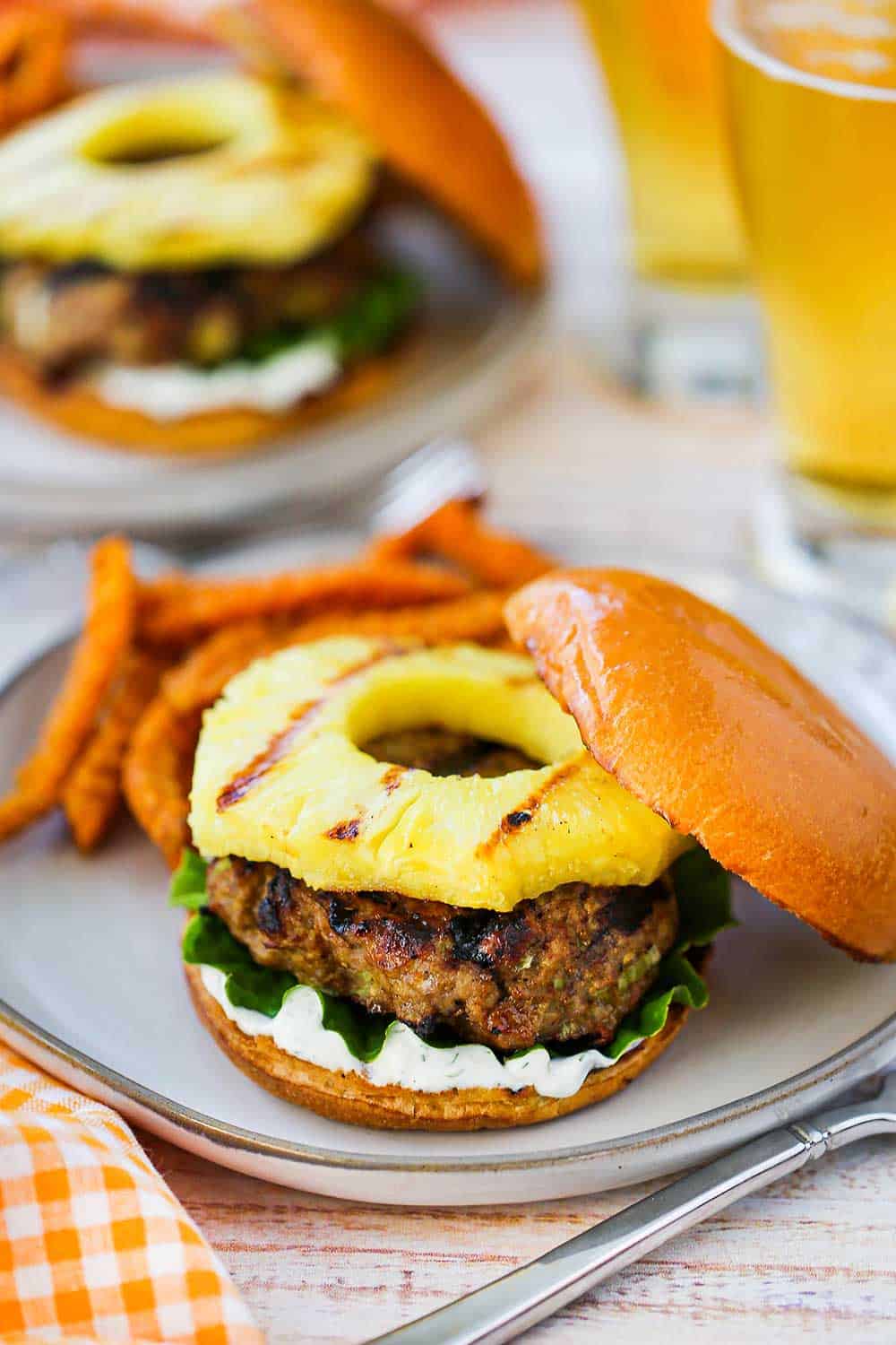 A curry turkey burger with grilled pineapple sitting on a plate next to sweet potato crinkle fries with a couple glasses of beer in the background.