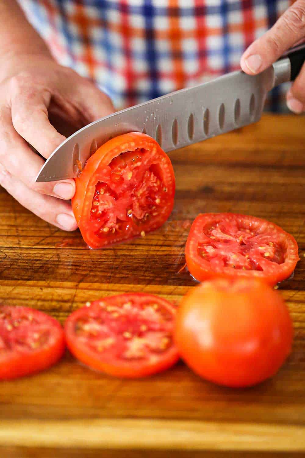 A person using a chef's knife to slice a very red tomato on a cutting board with other tomato slices nearby.