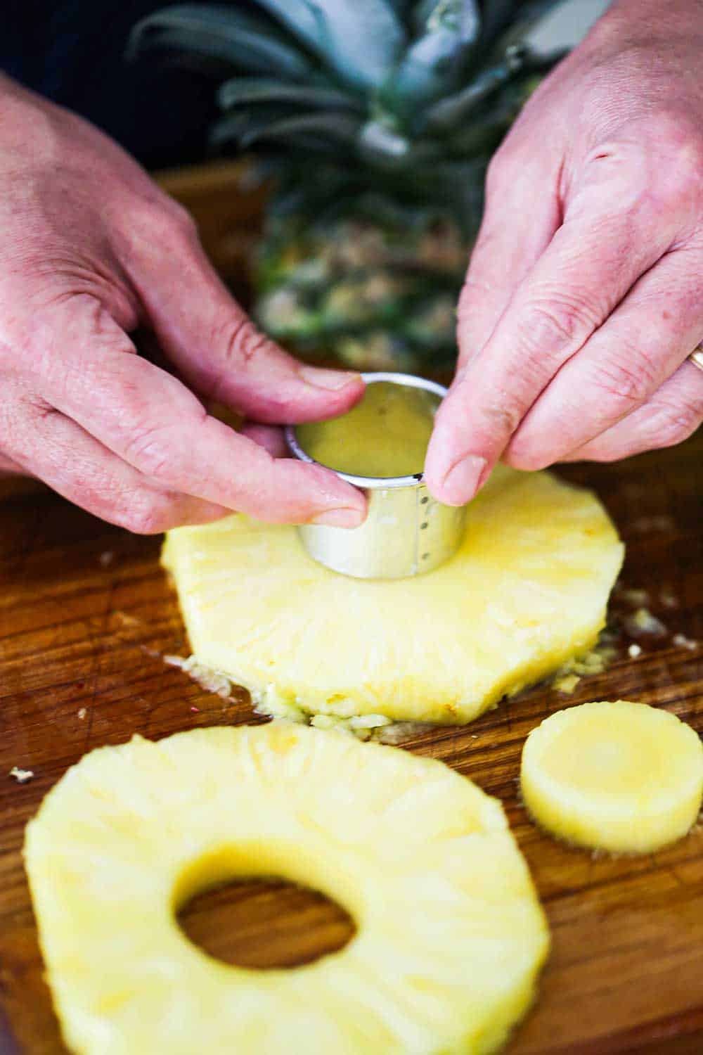 A pair of hands pressing a circular small cookie cutter into a pineapple slice on a wooden cutting board.