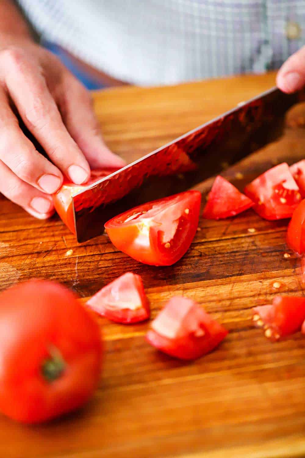 A person using a large Japanese chef's knife to slice a tomato into bite-sized pieces on a cutting board. 