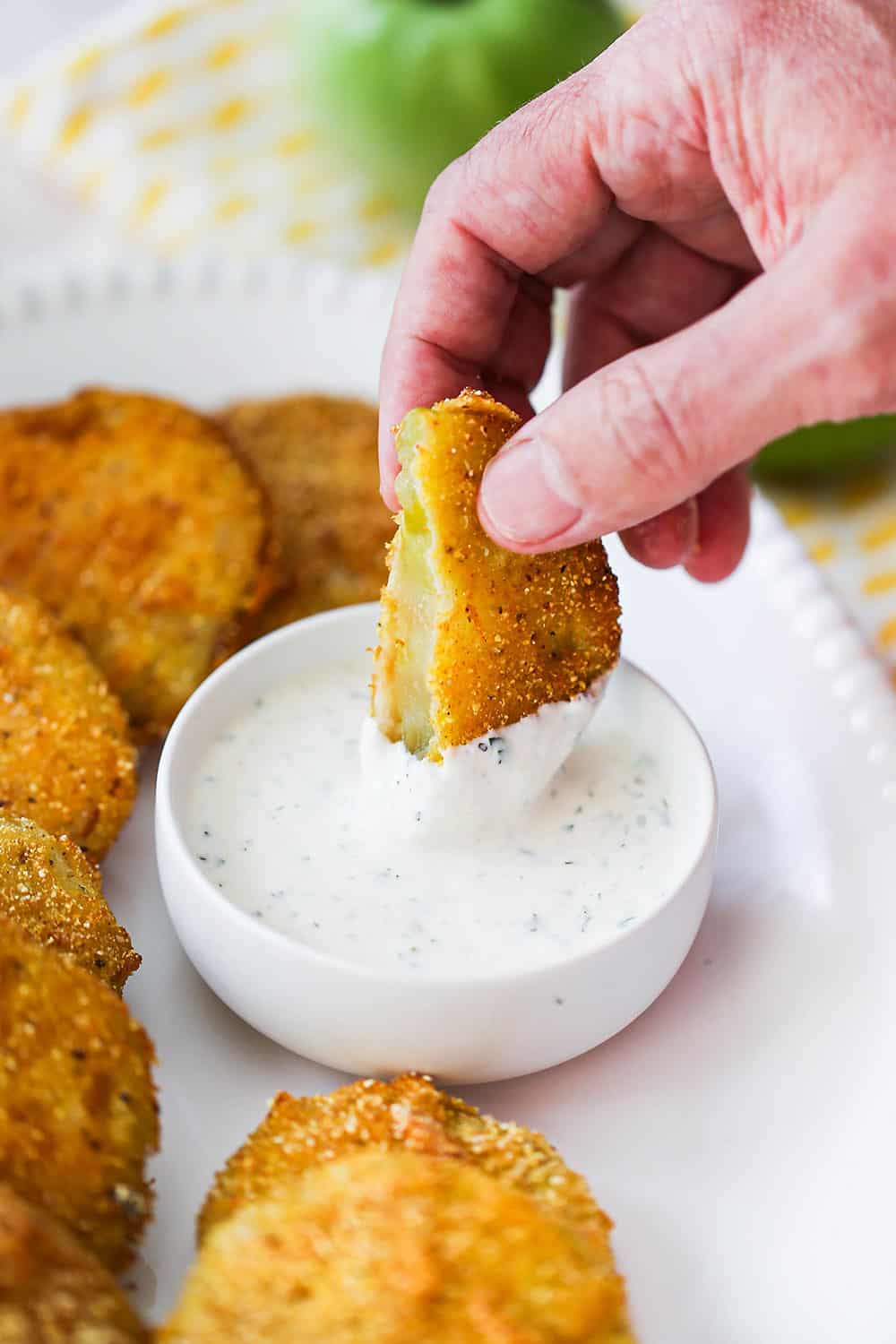 A hand dipping one half of a fried green tomato into a small white bowl of ranch dressing.