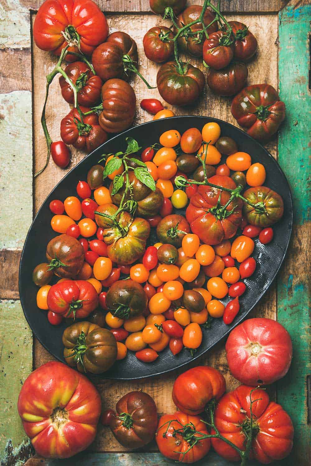 A bowl filled with and surrounded by a large variety of types of tomatoes including cherry, beefsteak, heirloom, and roma. 