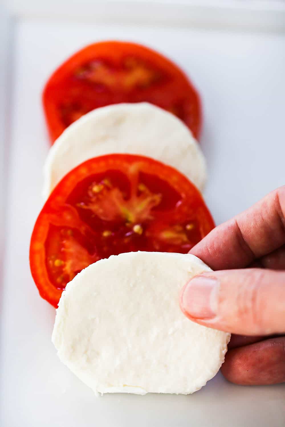 A person placing a circular slice of mozzarella cheese over a slice of tomato in front of two more layers of tomato and mozzarella.