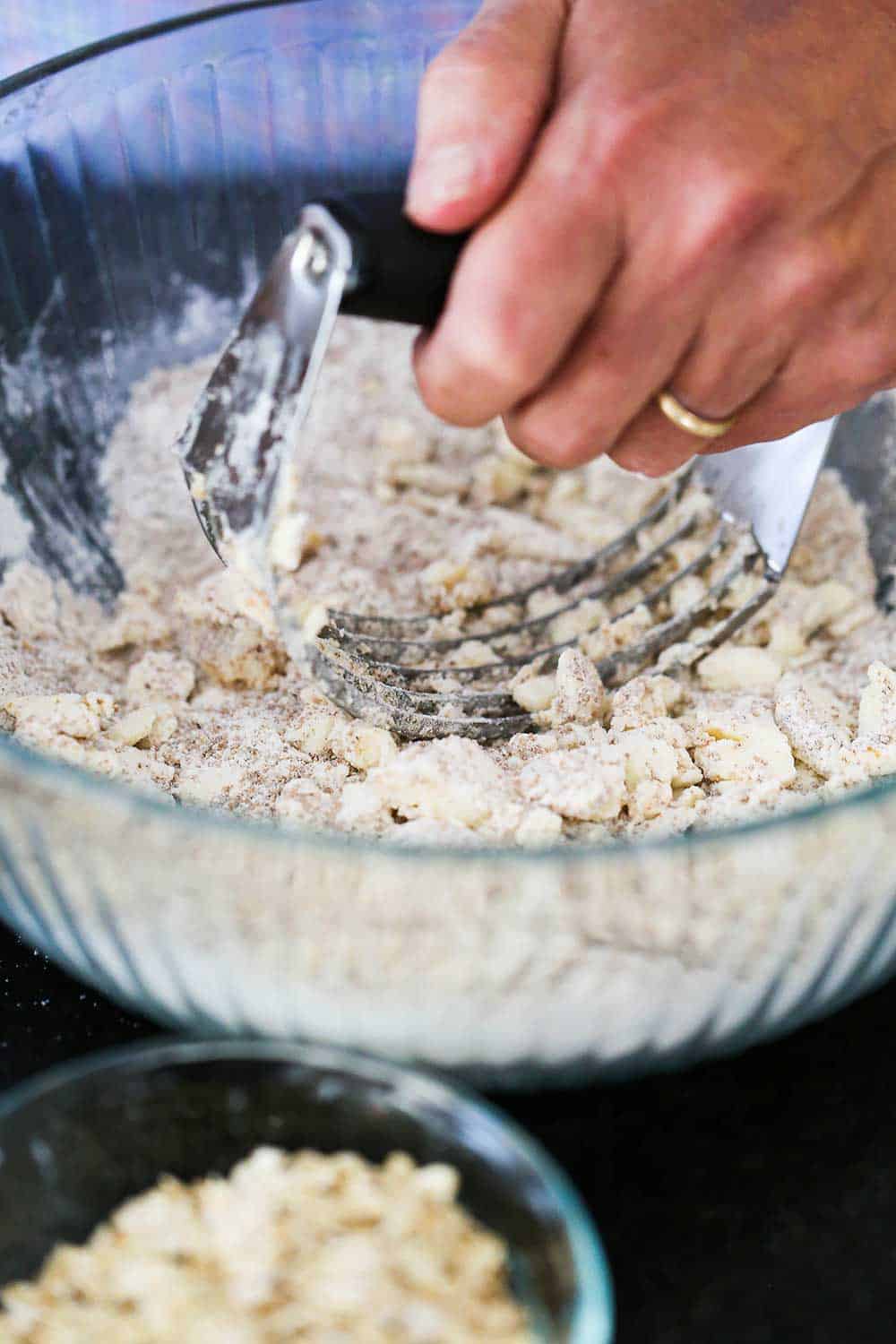 A person using a pastry blender to cut butter into a flour and brown sugar mixture in a large glass bowl.