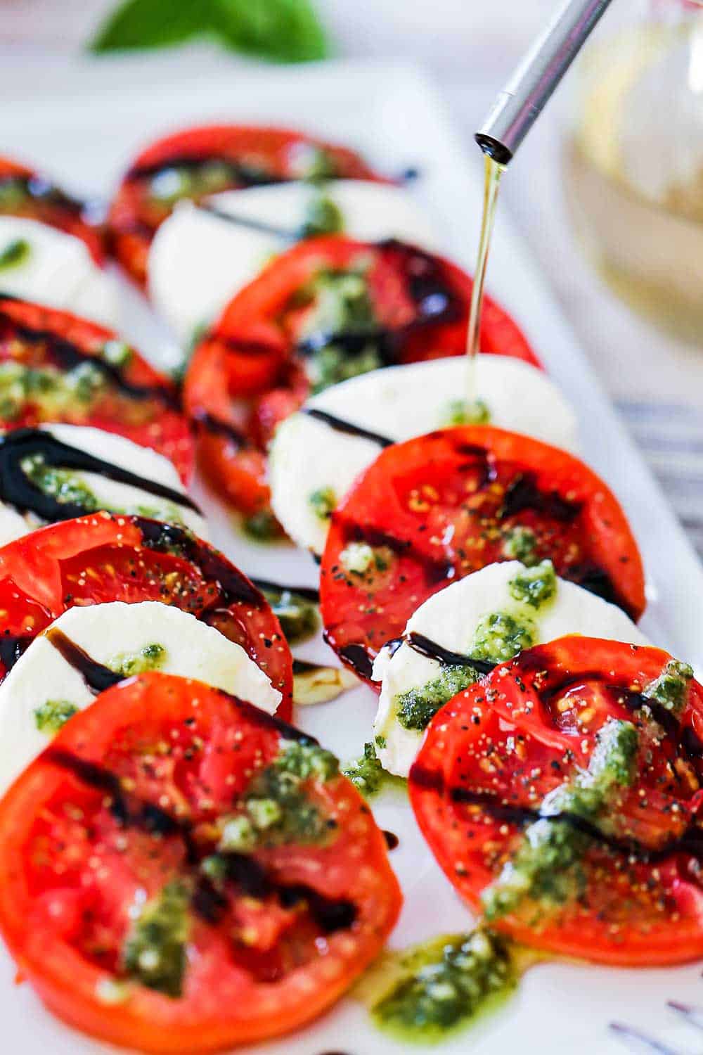 A thin drizzle of olive oil being poured from an olive oil dispenser onto a platter filled with caprese salad.