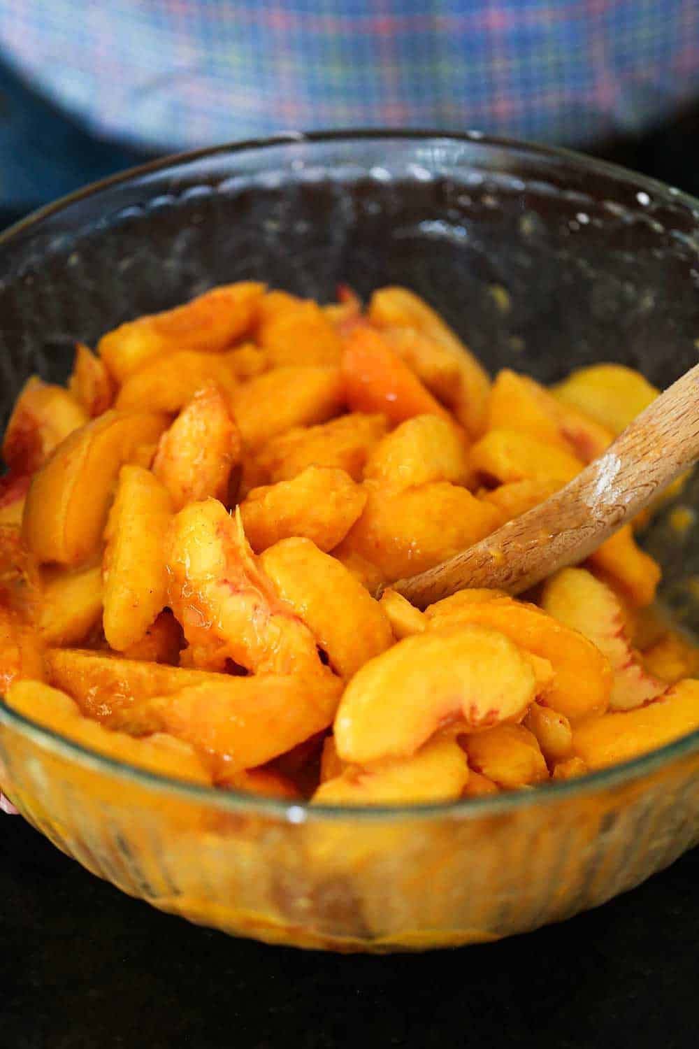 A large glass bowl filled with sliced fresh peaches with a wooden spoon inserted on the edge of the bowl.
