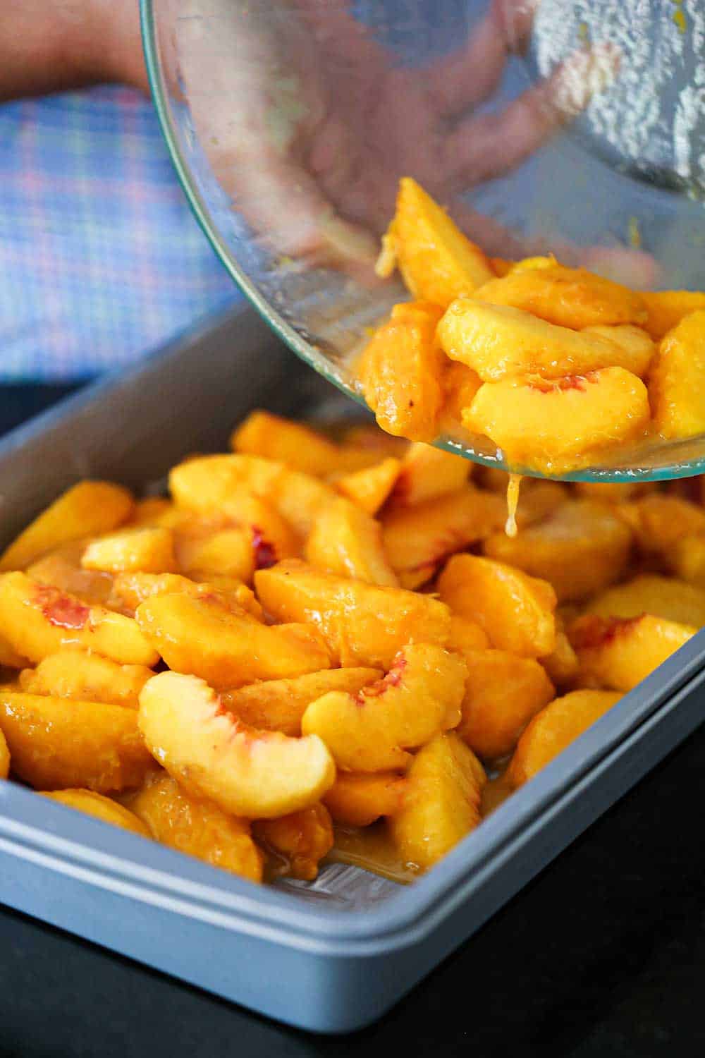 A person transferring sliced peaches in a sugar sauce from a glass bowl into a metal baking pan.