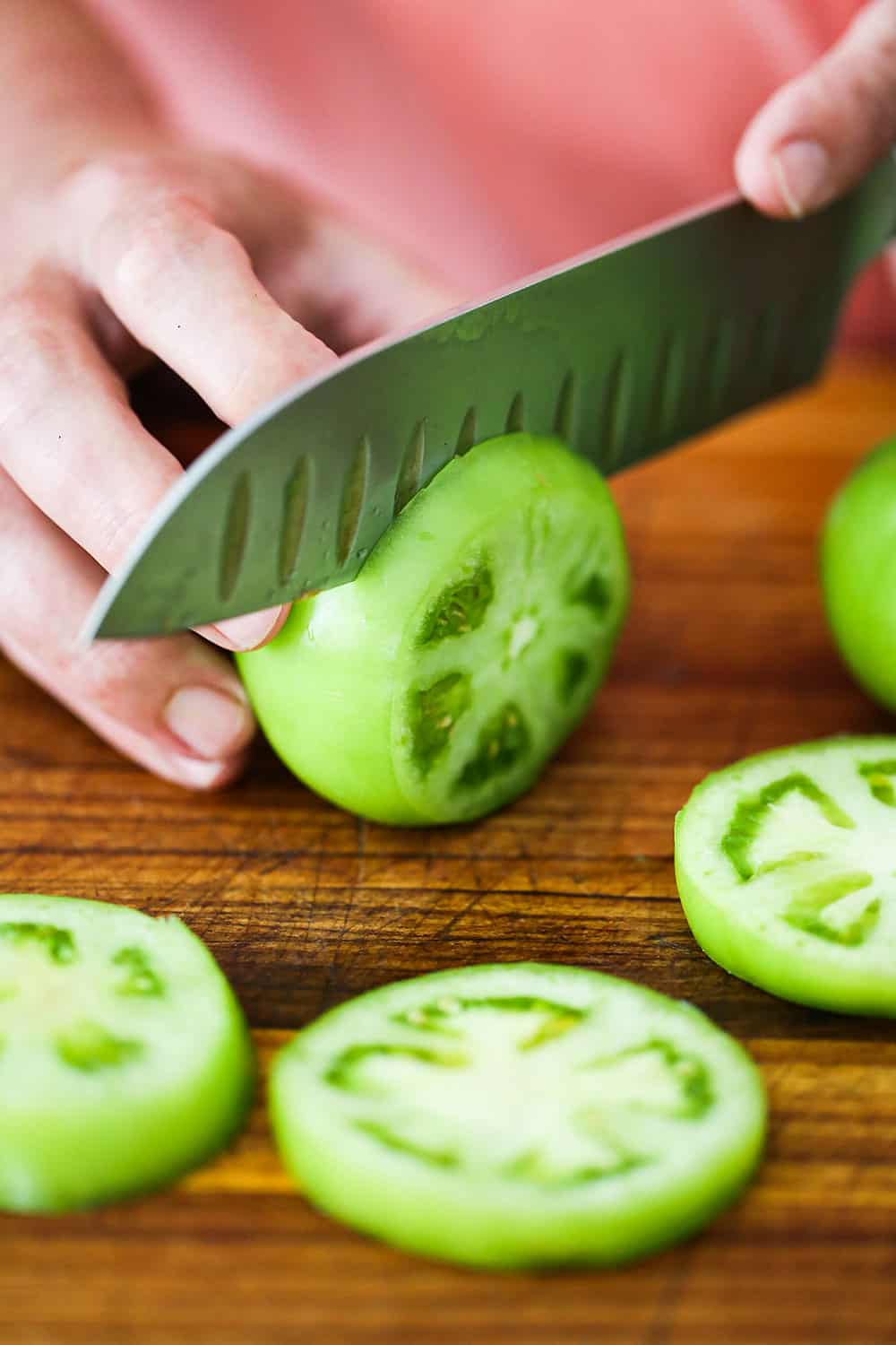 A person using a chef's knife to slice a green tomato on a cutting board with slices scattered around.