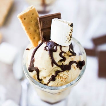 A glass vessel filled with s'more ice cream topped with a drizzle of chocolate sauce, graham crackers, chocolate candy, and a toasted marshmallow.