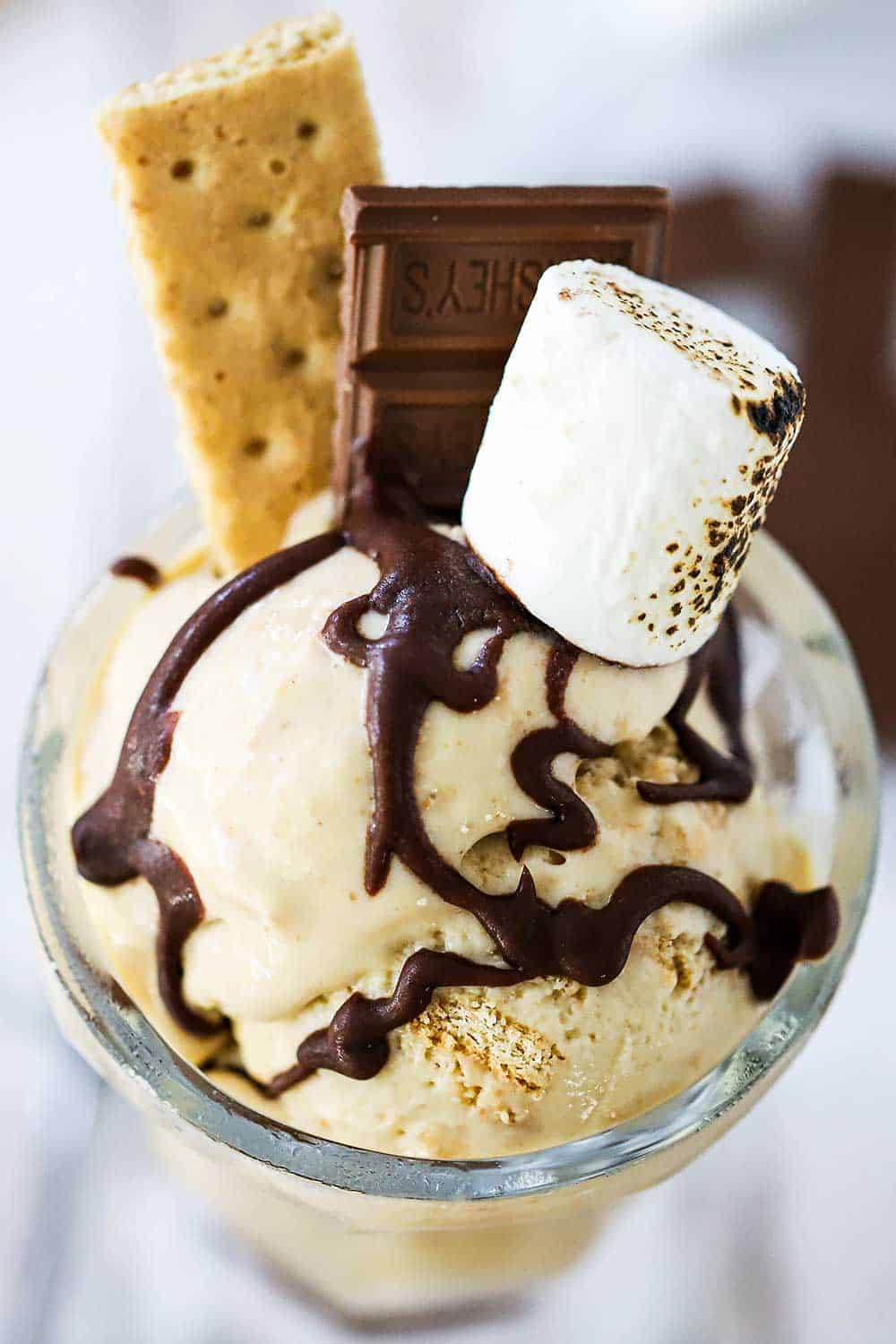 A glass vessel filled with s'more ice cream topped with a drizzle of chocolate sauce, graham crackers, chocolate candy, and a toasted marshmallow.