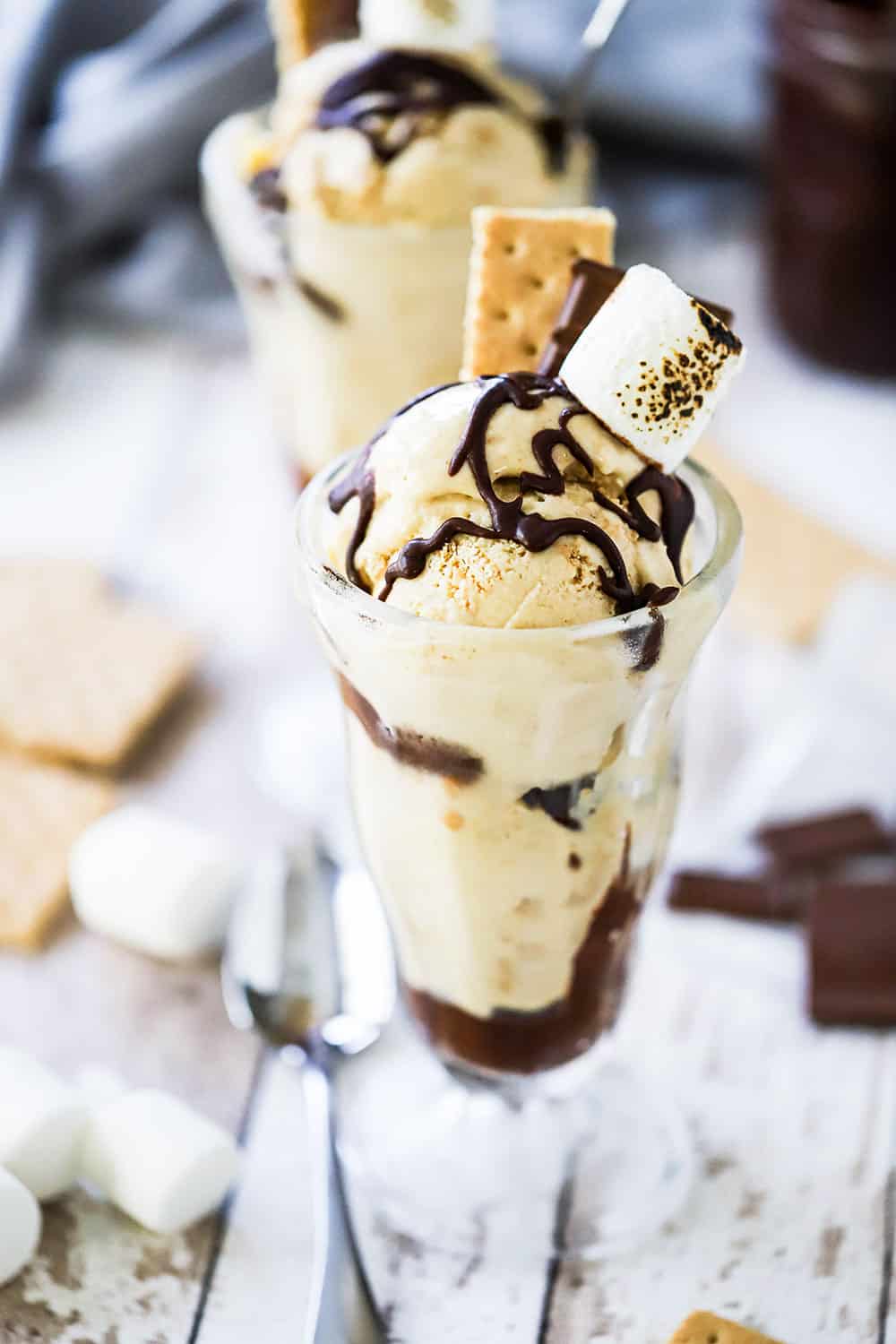 An ice cream parlor glass holder filled with s'mores ice cream topped with a piece of graham cracker, a piece of chocolate and a toasted marshmallow.