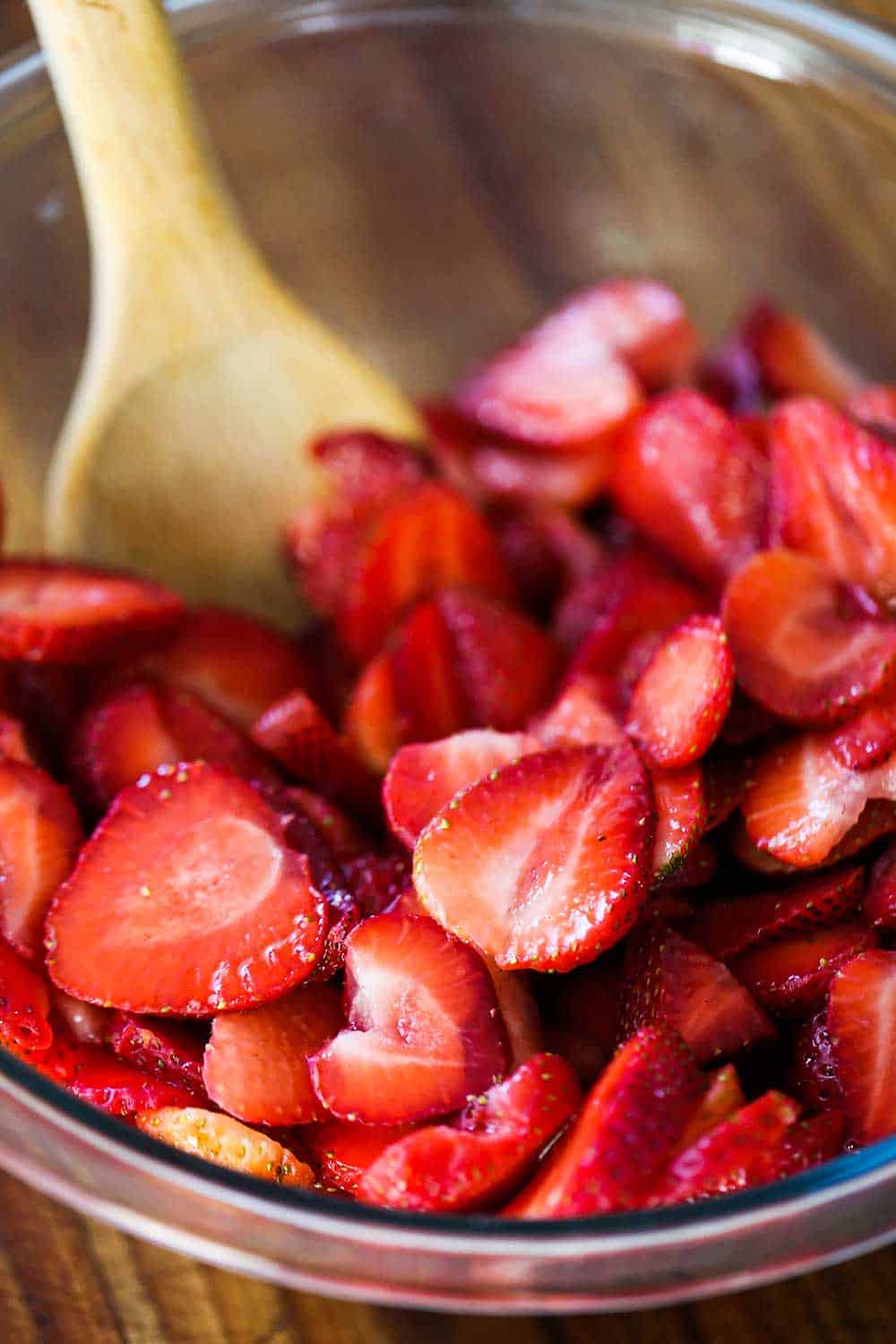 A large wooden spoon stirring sliced strawberries coated in sugar all in a large glass bowl.