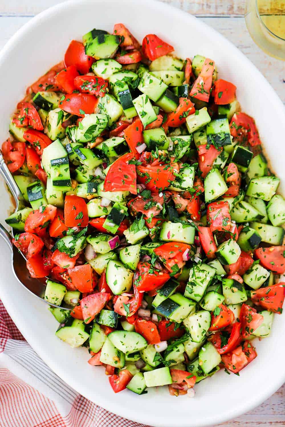 A large white oval serving dish filled with a cucumber tomato salad next to a red checkered napkin. 