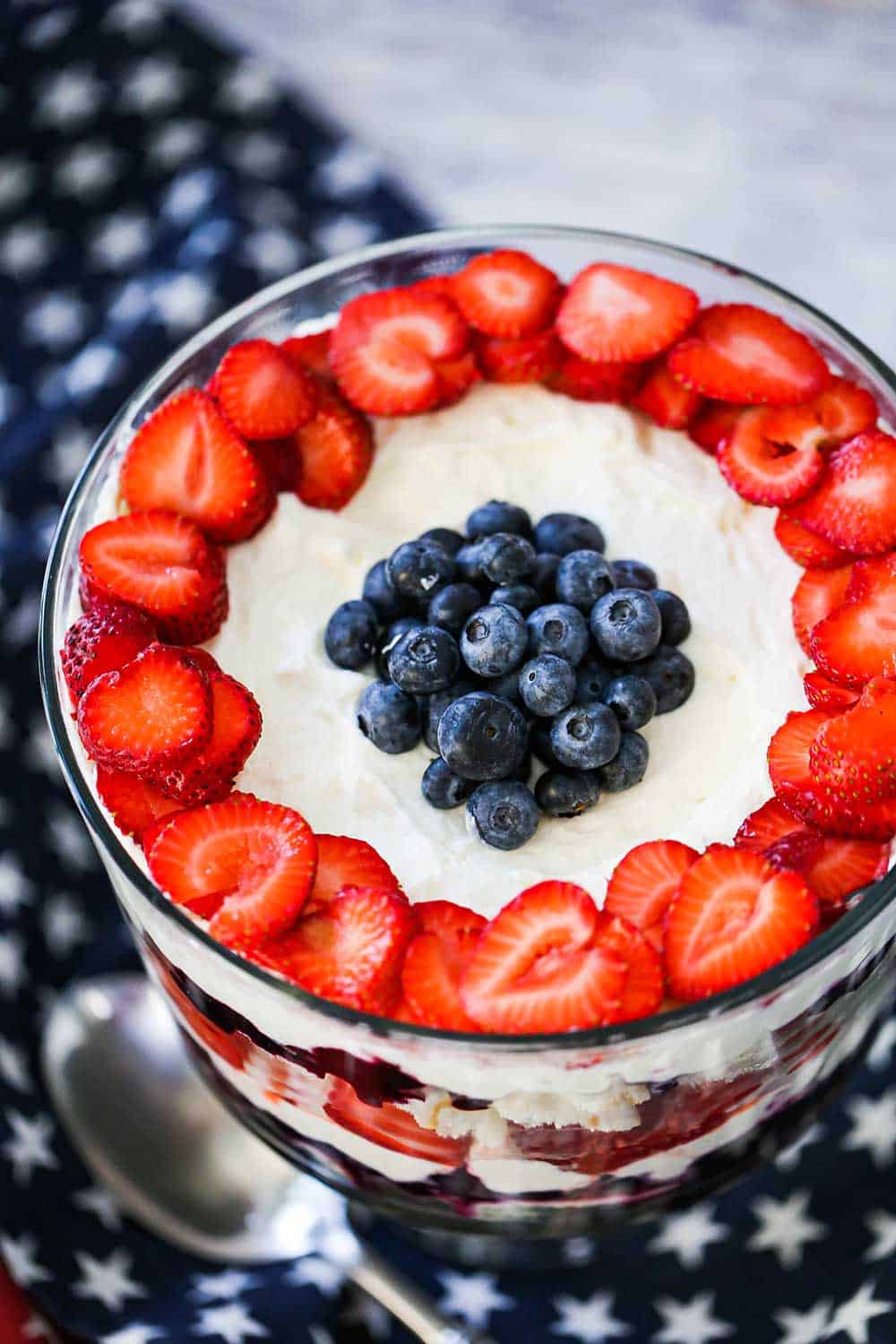 The top of a red, white, and blue trifle that is garnished with an outer circle of fresh, sliced strawberries, and then a pile of fresh blueberries in the center, all sitting on white cream sauce.