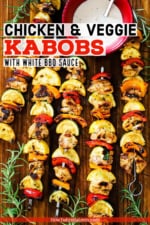 Five grilled chicken kabobs on metal skewers lined with vegetables and a red bowl of white BBQ sauce nearby.