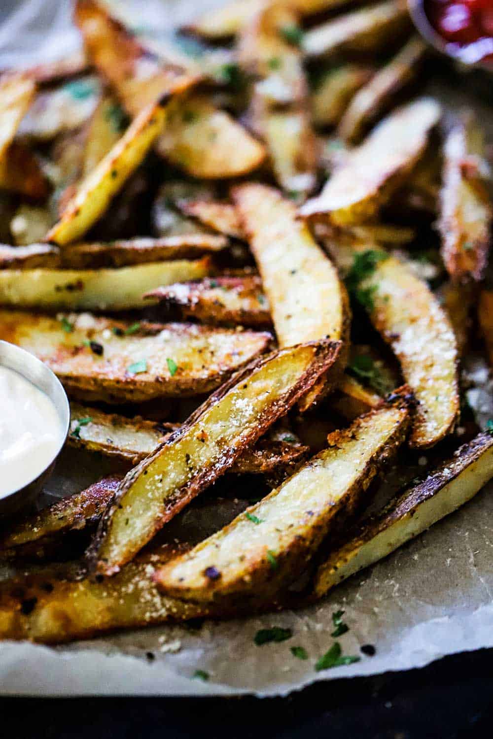A baking sheet lined with parchment paper with layers of garlic parmesan steak fries sitting next to a small dipping bowl filled with aioli.