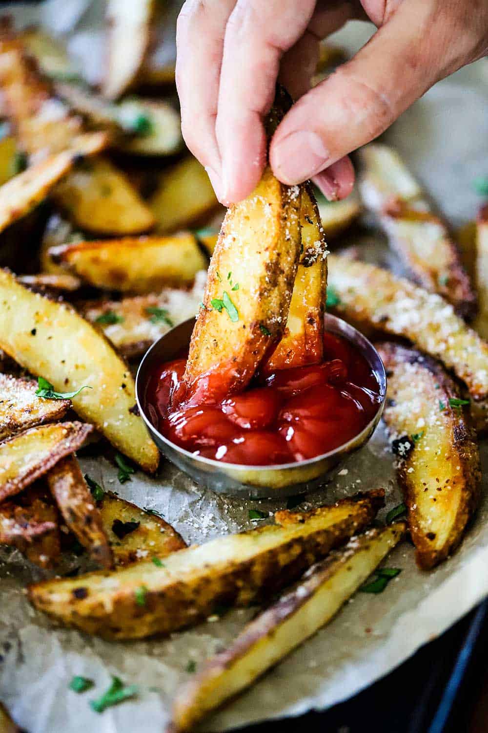 A person using his fingers to plunge to oven-roasted steak fries into a metal condiment holder filled with ketchup, all on a pan will with other fries.
