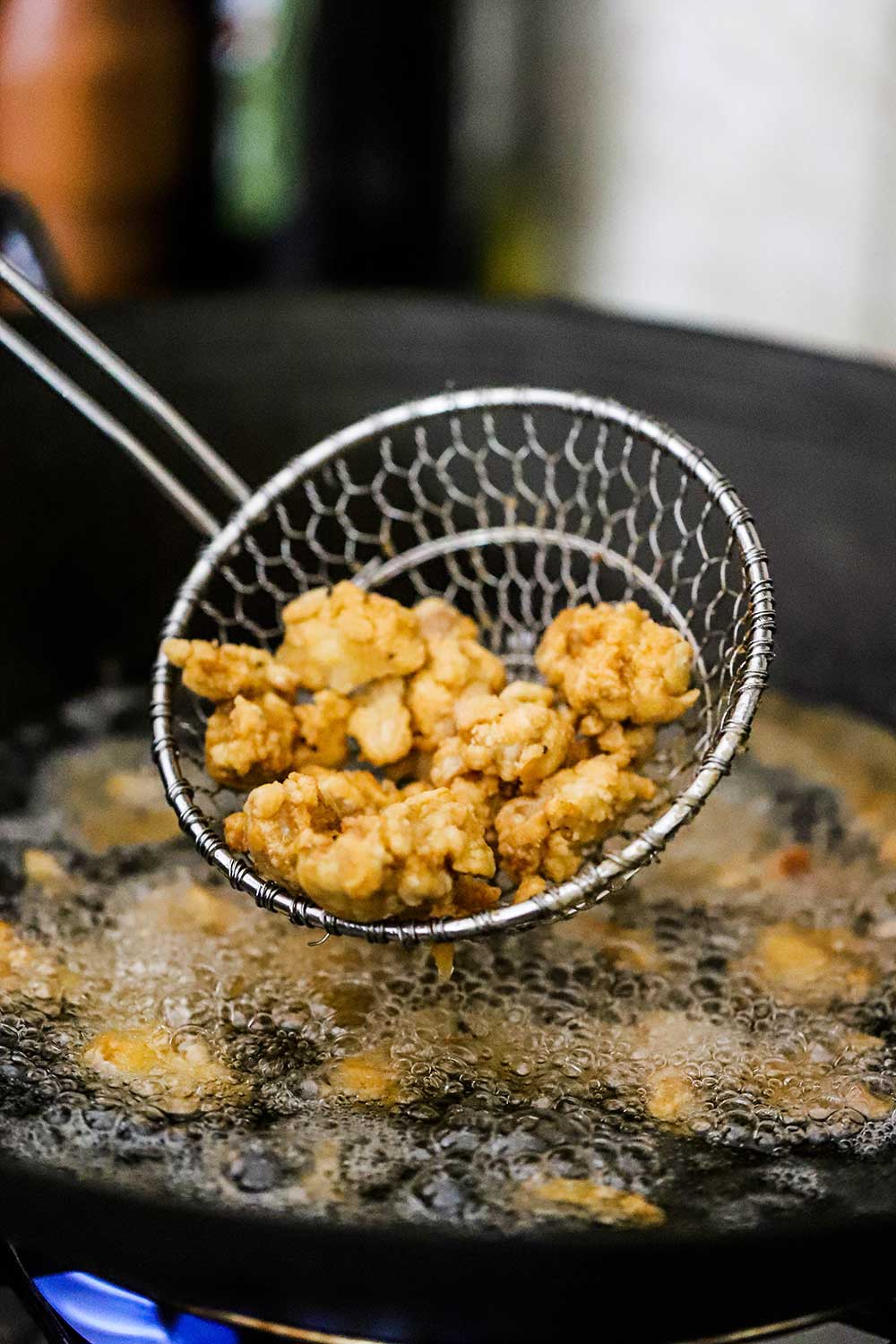 A kitchen spider holding piece of fried chicken over a wok filled with hot oil.