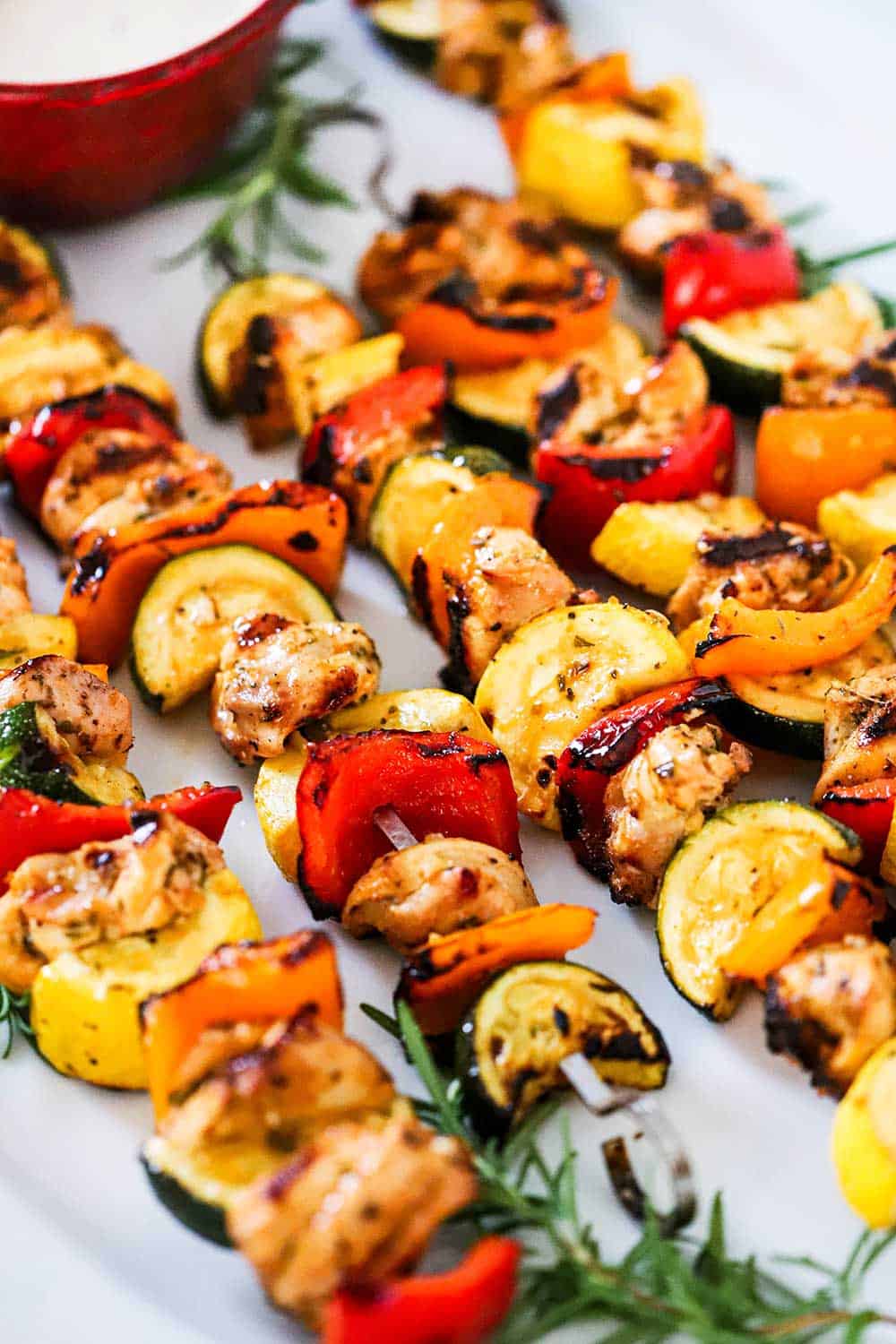 A row of 5 chicken kabobs on a white oval platter garnish with fresh rosemary sprigs and a bowl of white sauce. 