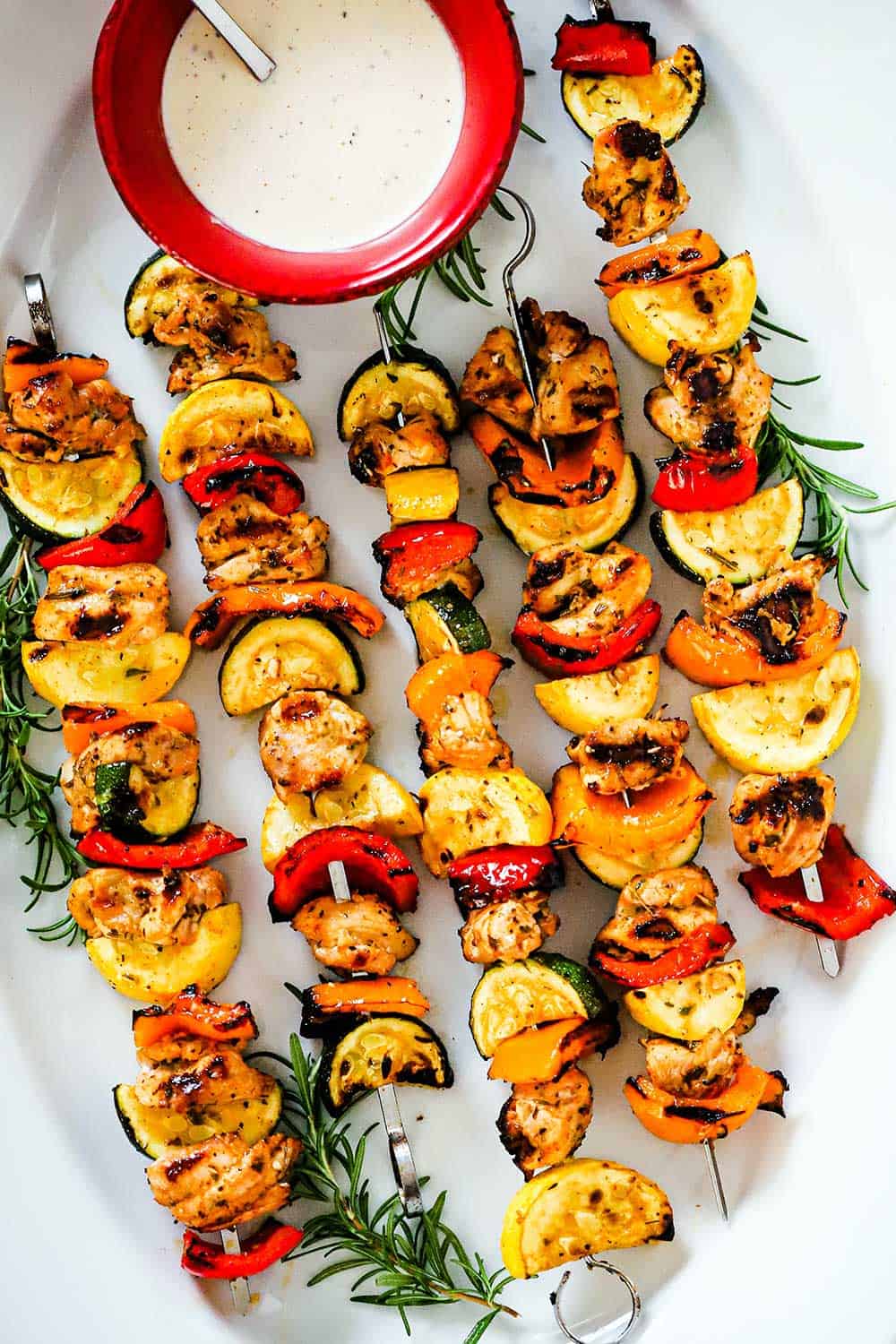 A large white oval platter filled with grilled chicken and vegetable kabobs with a red bowl filled with white BBQ sauce nestled in. 