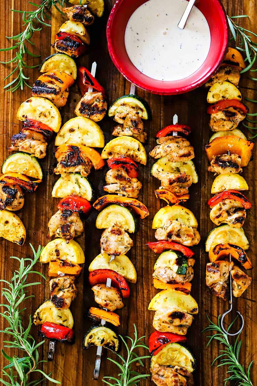 Five grilled chicken kabobs on metal skewers lined with vegetables and a red bowl of white BBQ sauce nearby.