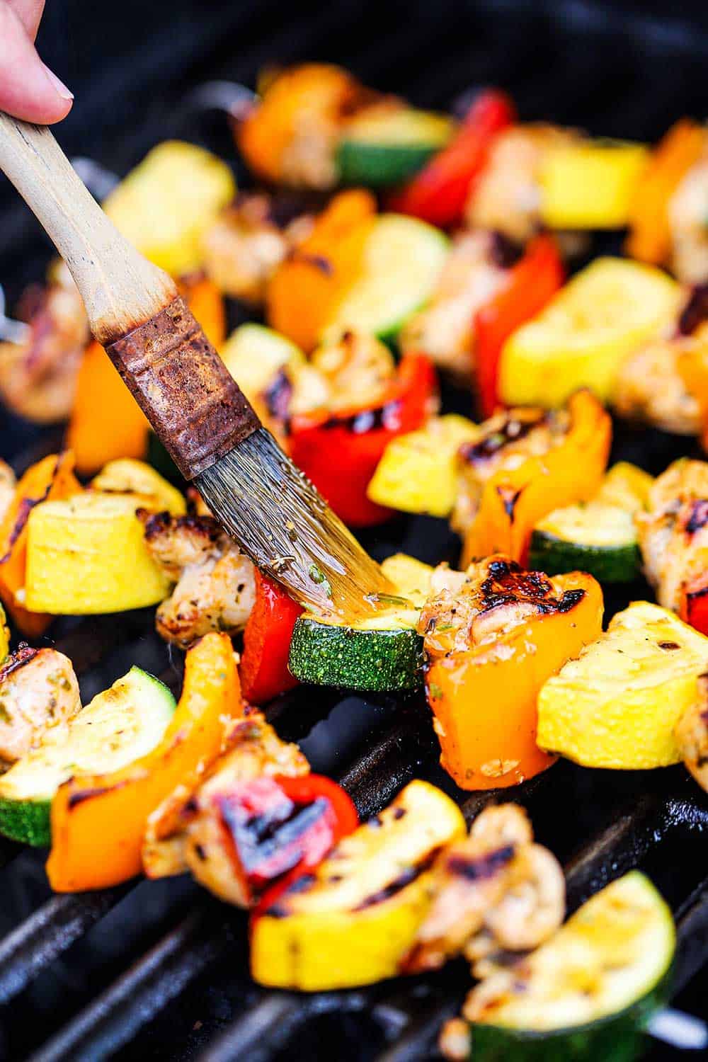 A person brushing marinade onto skewers on a grill that contain pieces of uncooked chicken as well as bell peppers, squash, and zucchini pieces. 