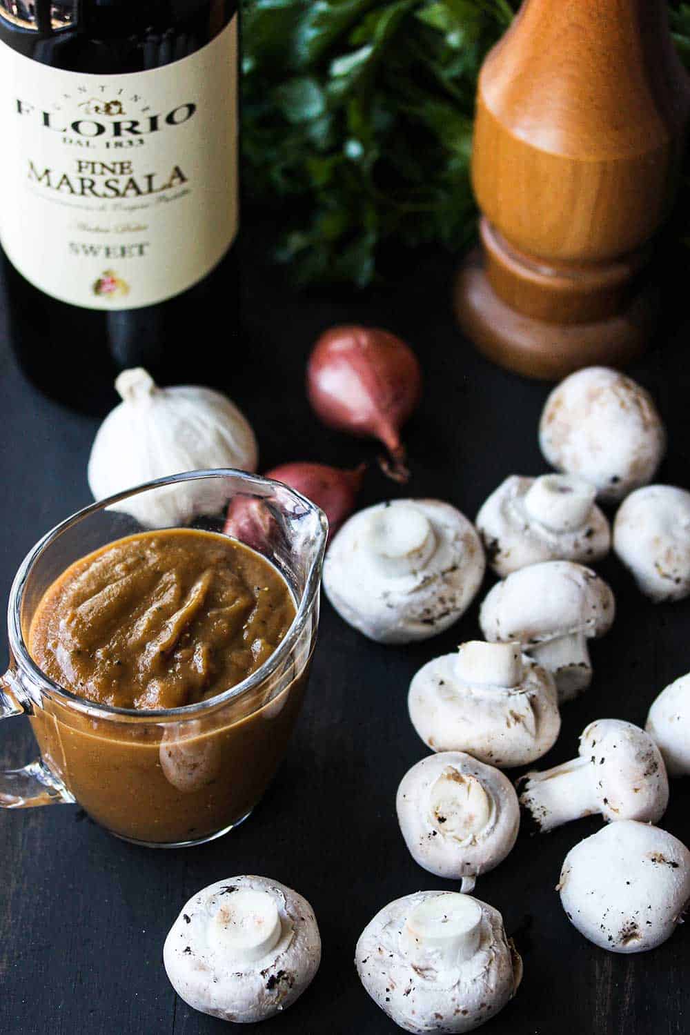 A wooden cutting board with whole button mushrooms, a small glass pitcher of brown sauce, whole garlic, shallots, a bottle of marsala wine, and a pepper grinder on it.