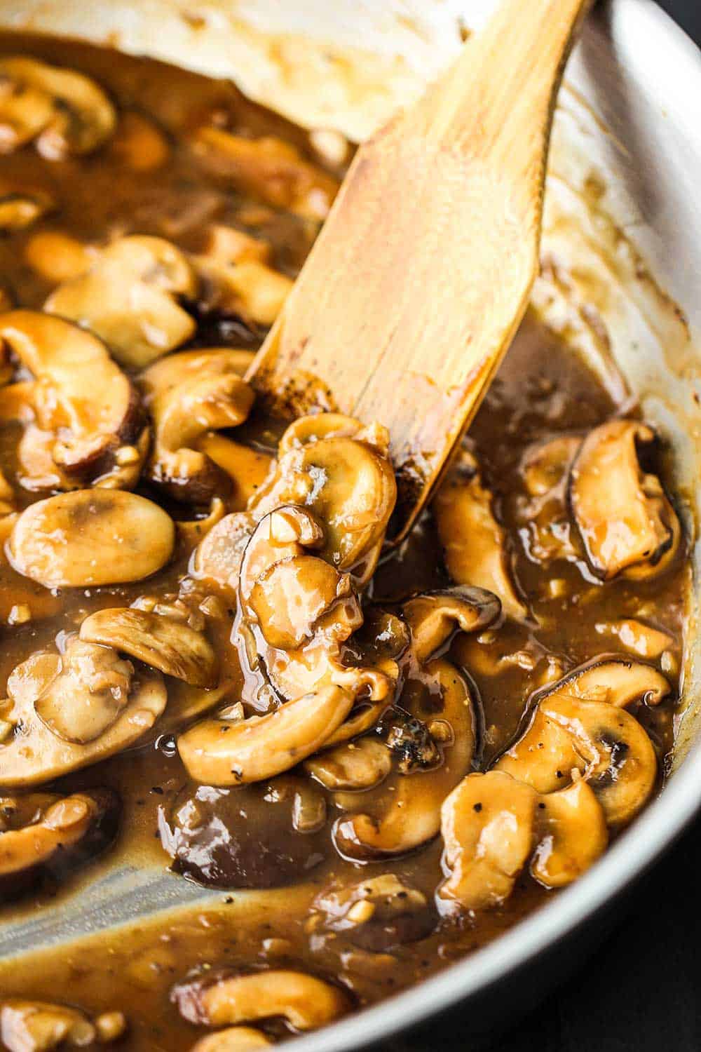 A stainless steel skillet filled with a marsala sauce and sliced mushrooms with a wooden spatula off to the side.