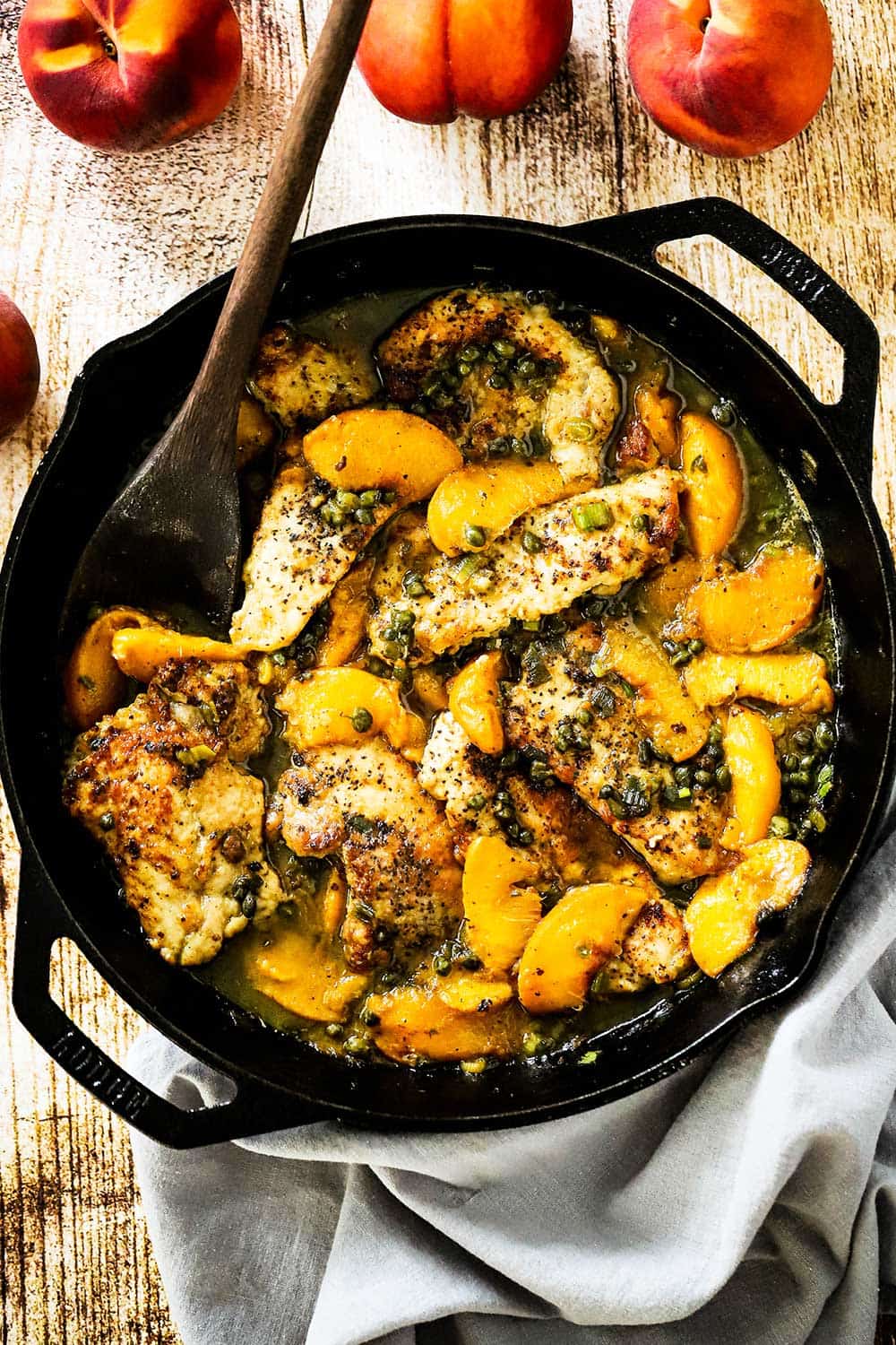 A large black cast-iron skillet filled with pan-fried pork cutlets and peaches in a savory sauce all next to several whole peaches.