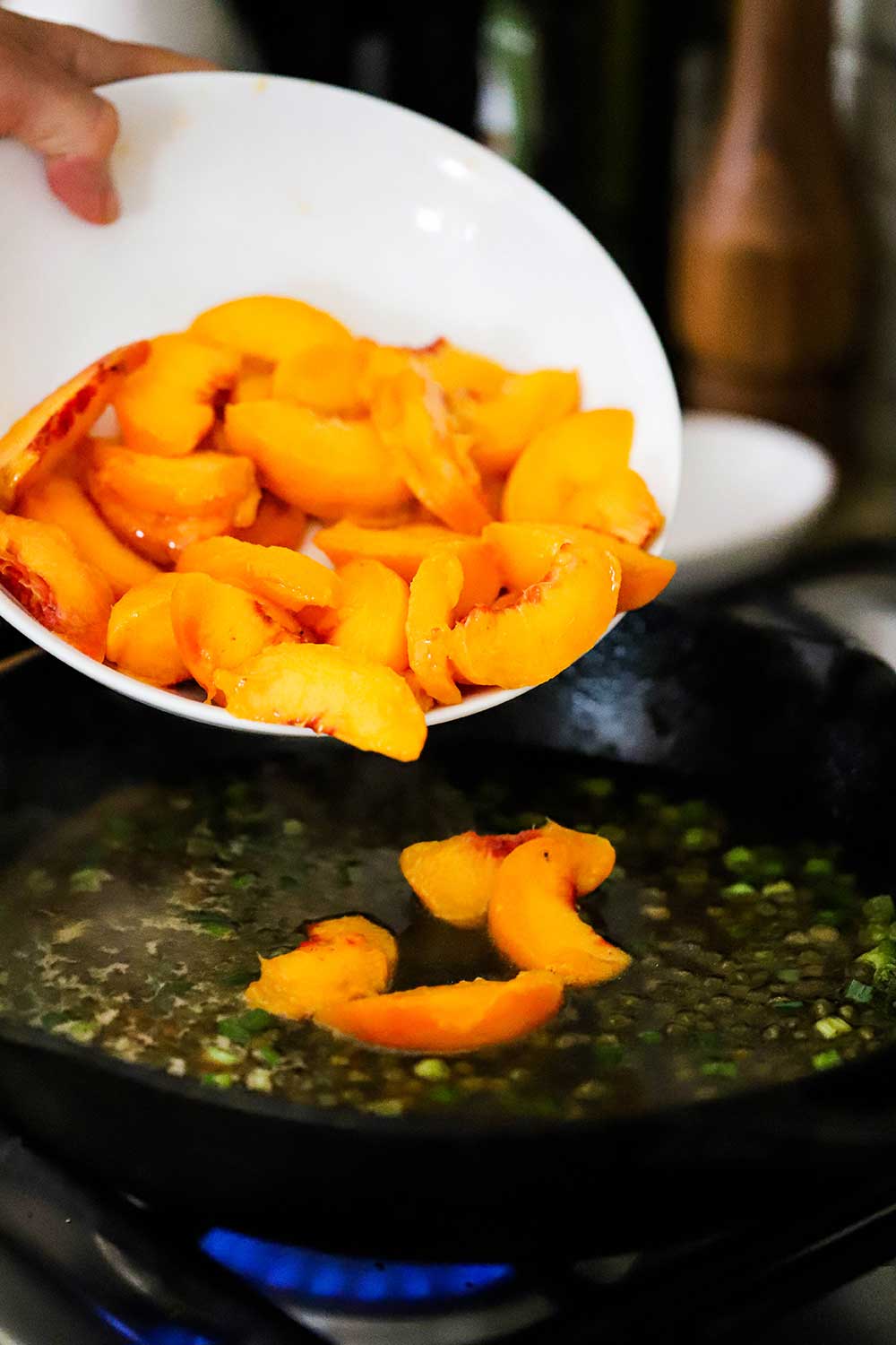 A person transferring sliced fresh peaches into a cast-iron skillet with a simmering sauce with capers, scallions, and chicken stock. 