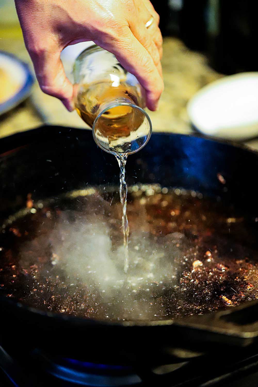 A person pouring sherry from a small carafe into a cast-iron skillet with pieces of breading stuck to the surface of the pan. 
