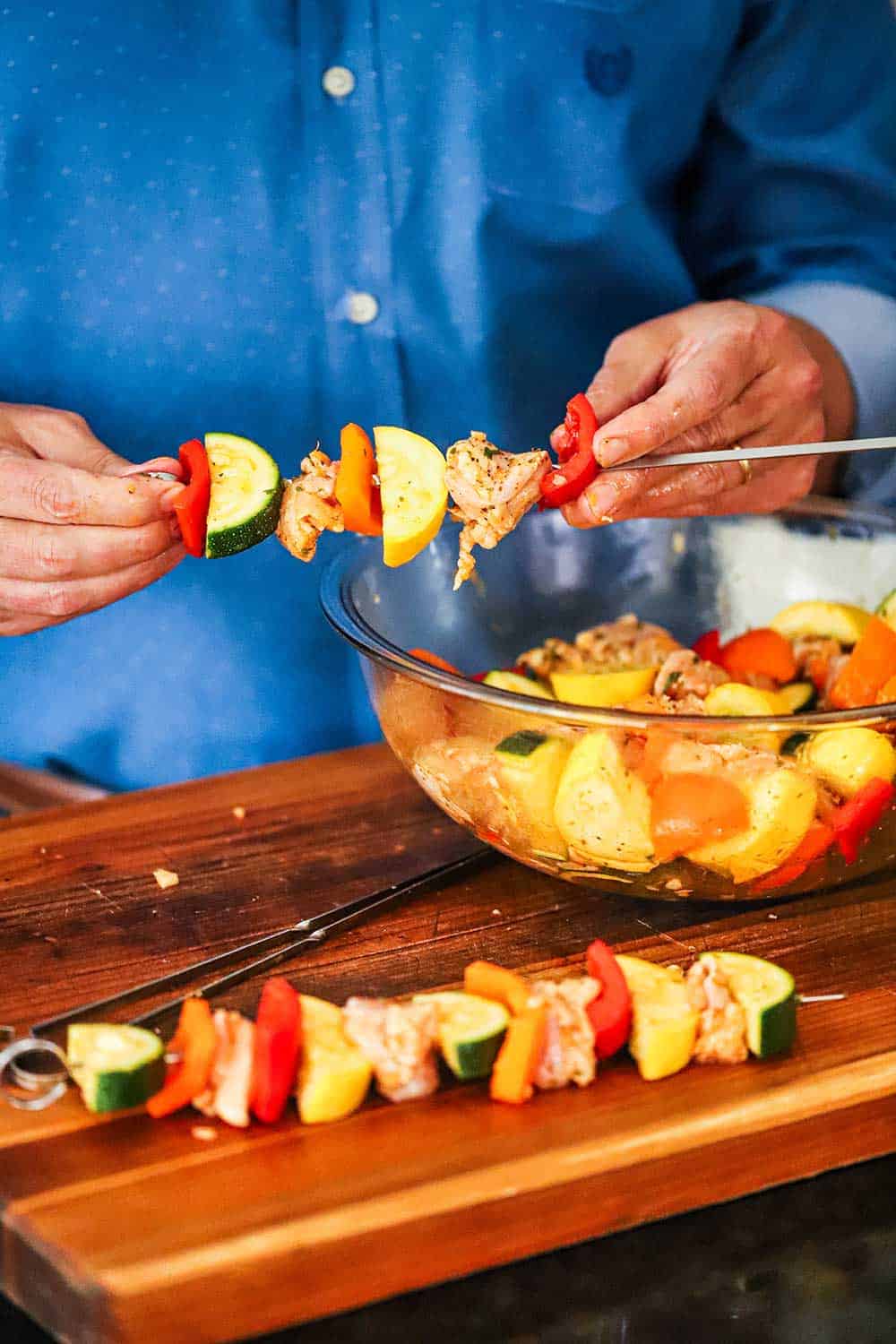 A person in a blue shirt adding pieces of cut chicken and vegetables onto metal skewers. 