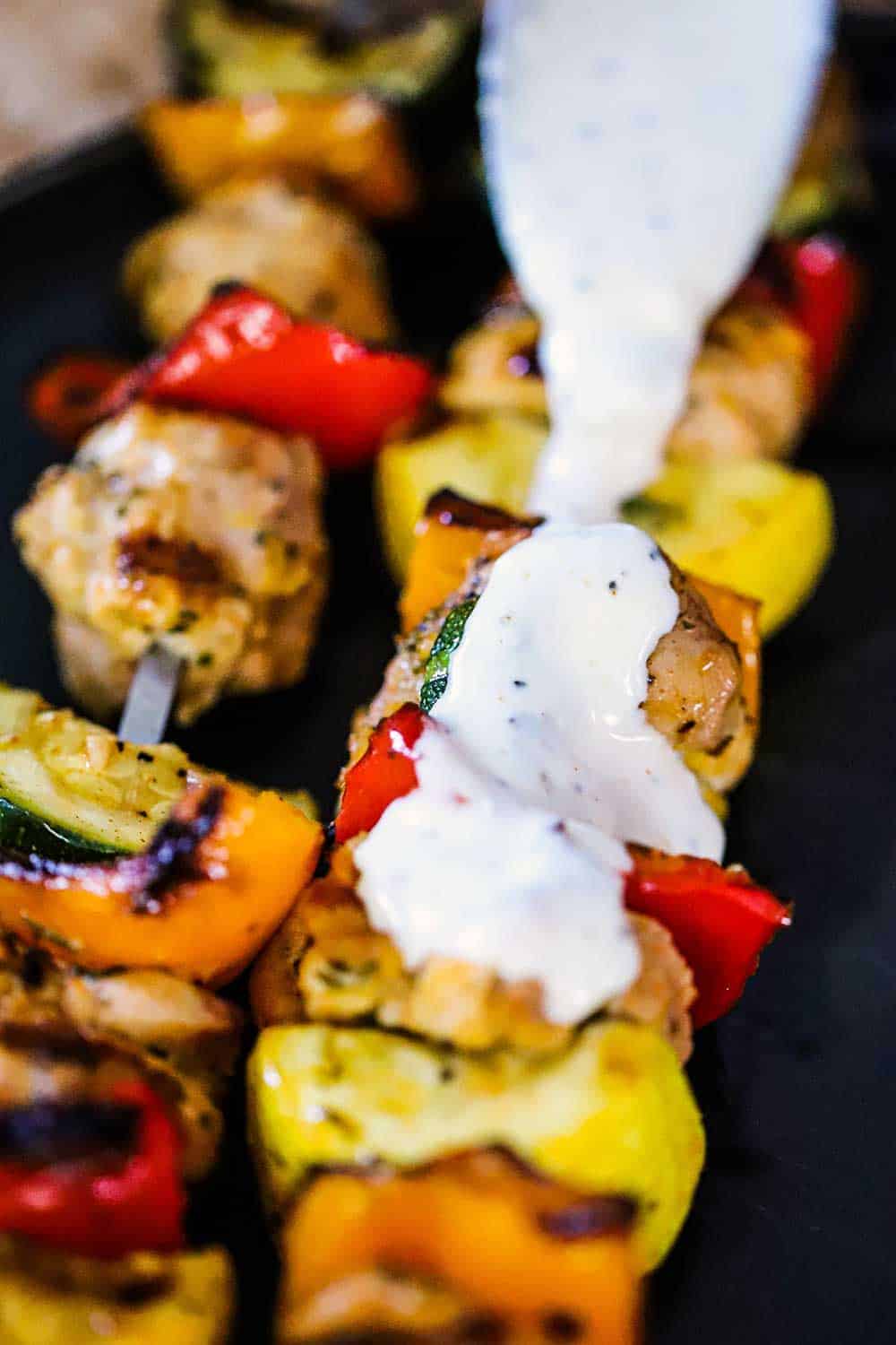 A spoon drizzling white BBQ sauce over a grilled chicken and vegetable kabob. 