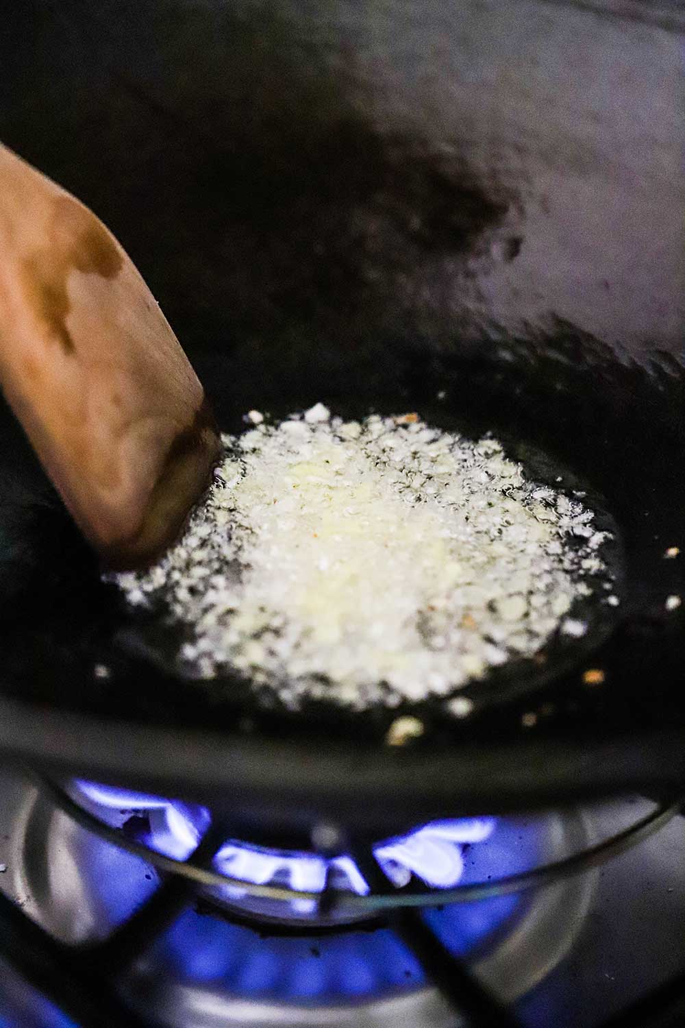 A wok on a gas-burning stove with hot oil and simmering minced garlic and ginger in it.