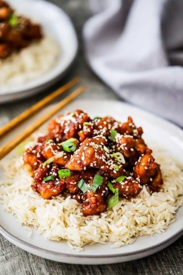 A dinner plate filled with a bed of white rice with sesame chicken on top of it and a pair of chop sticks nearby.