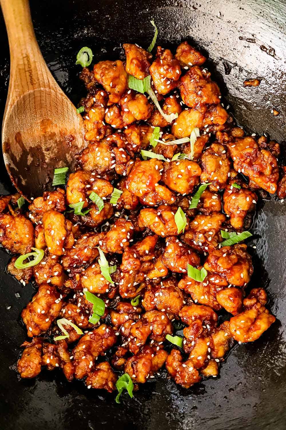 A wok filled with sesame chicken with slivers of scallions scattered on top and a wooden spoon off to the side.