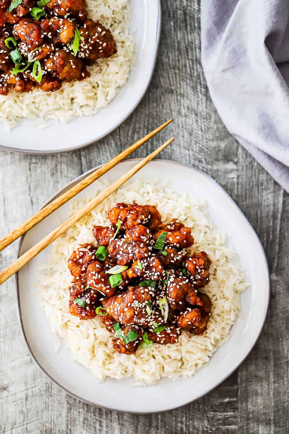 Two dinner plates both filled with a bed of white rice with sesame chicken on top of it and a pair of chop sticks nearby.