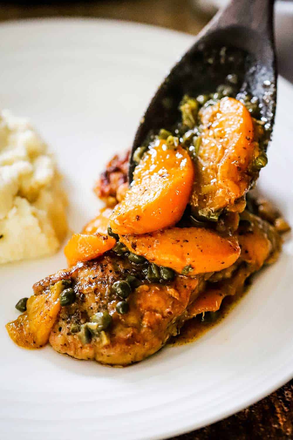 A wooden spoon transferring sautéed peaches and capers onto pan-fried pork cutlets on a white dinner plate.