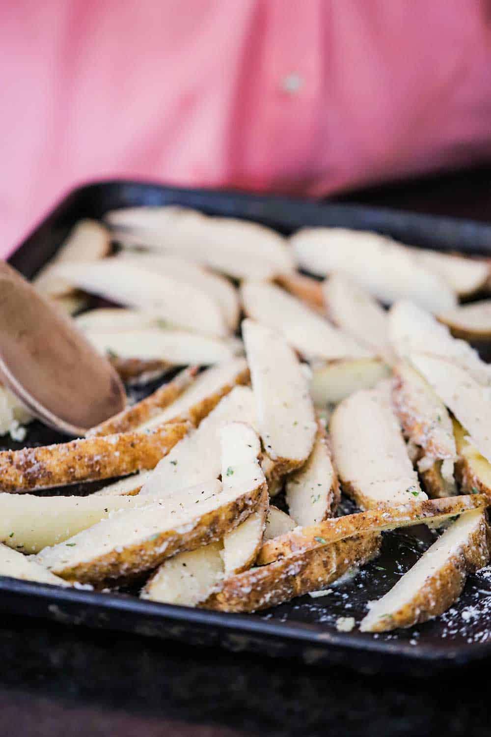 A person using a wooden spoon to spread potato wedges covered with cheese and herbs on a baking sheet.