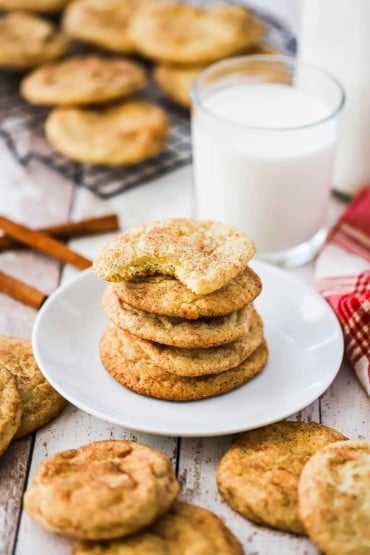 A stack of snickerdoodle cookies on a small white plate surrounded by other cookies and a glass of milk.