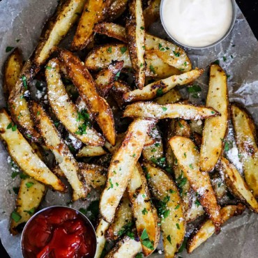 A pile of garlic parmesan steak fries sitting on a board lined with parchment paper with a small bowl of ketchup on one side and a bowl of aioli on the other side.