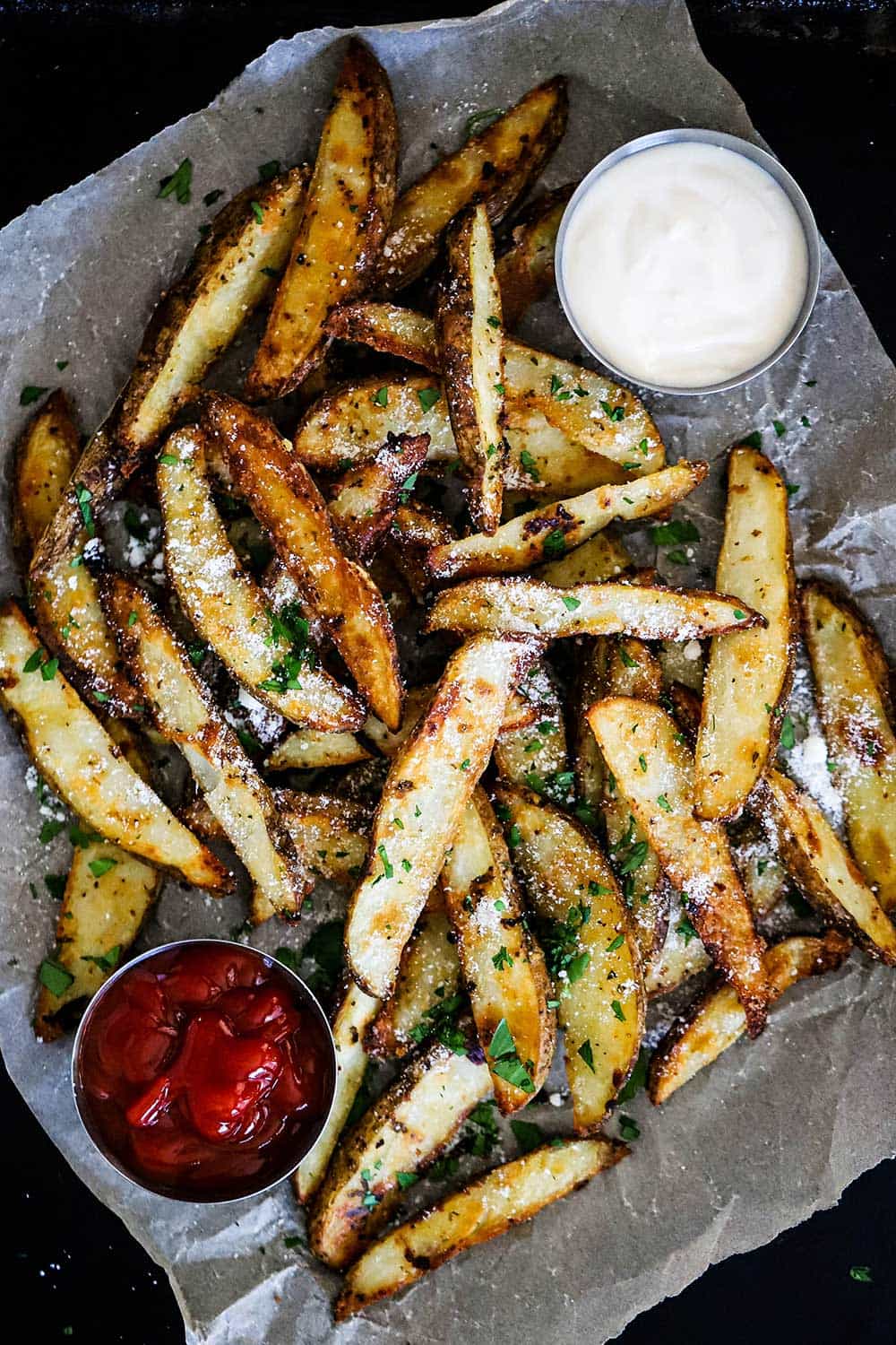 A pile of garlic parmesan steak fries sitting on a board lined with parchment paper with a small bowl of ketchup on one side and a bowl of aioli on the other side.