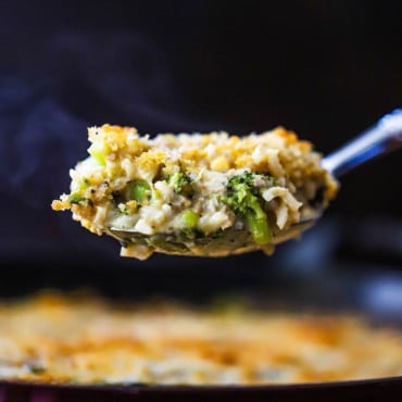 A large silver spoon lifting up a steaming helping of broccoli casserole over a dish of the same.