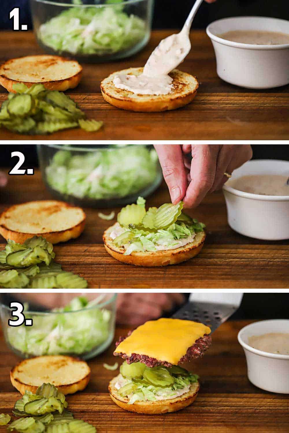 A person spooning Russian dressing onto a bottom hamburger bun and then that person placing lettuce and pickles on it and then a hamburger patty with cheese on top.